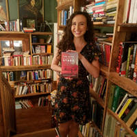 Francesca Varela holding her book, The Seas of Distant Stars, standing in front of bookshelves at a bookstore in Portland, Oregon.